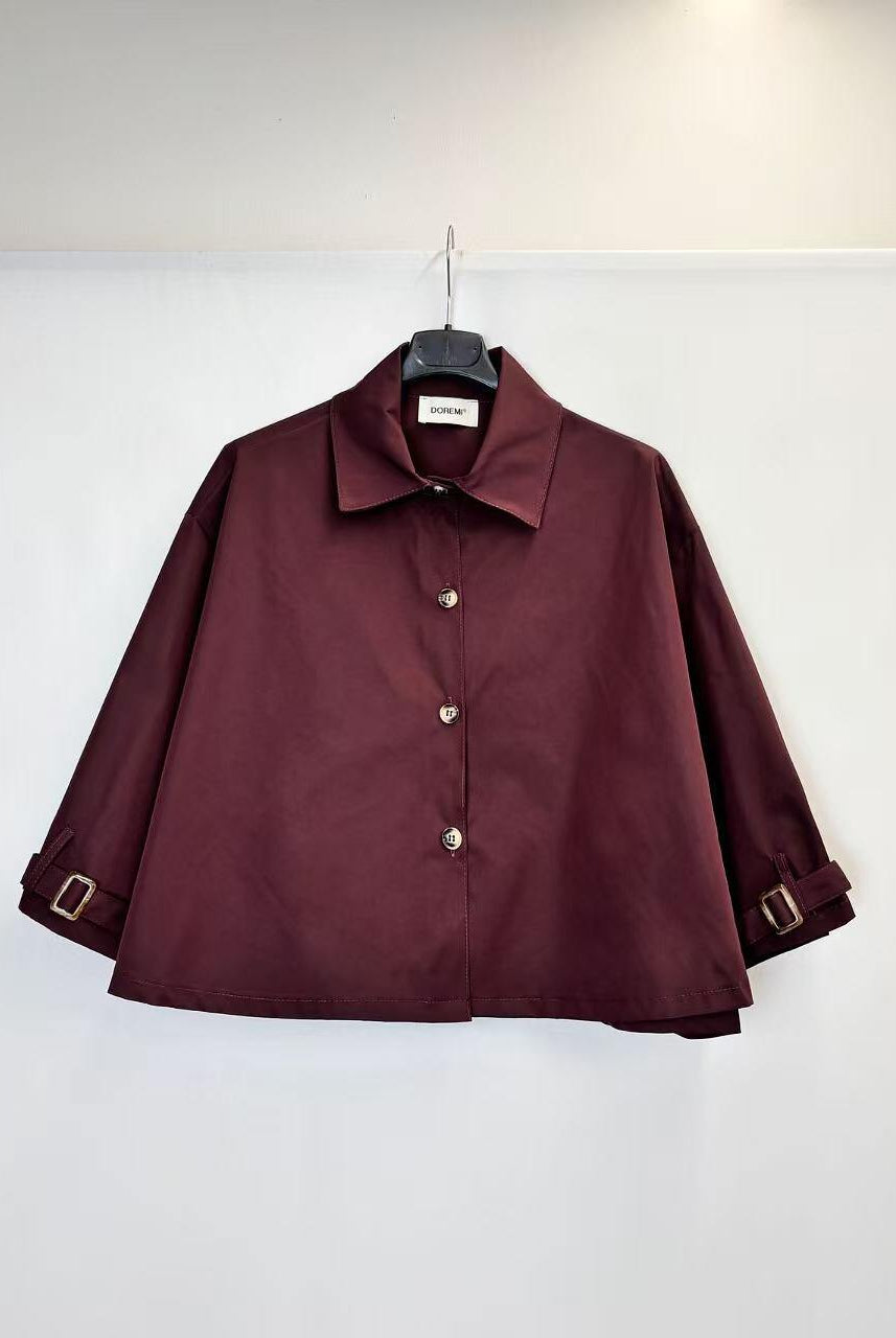 A dark burgundy long-sleeve trench jacket with a collar and decorative silver-tone buttons, hanging on a black hanger against a plain white wall.