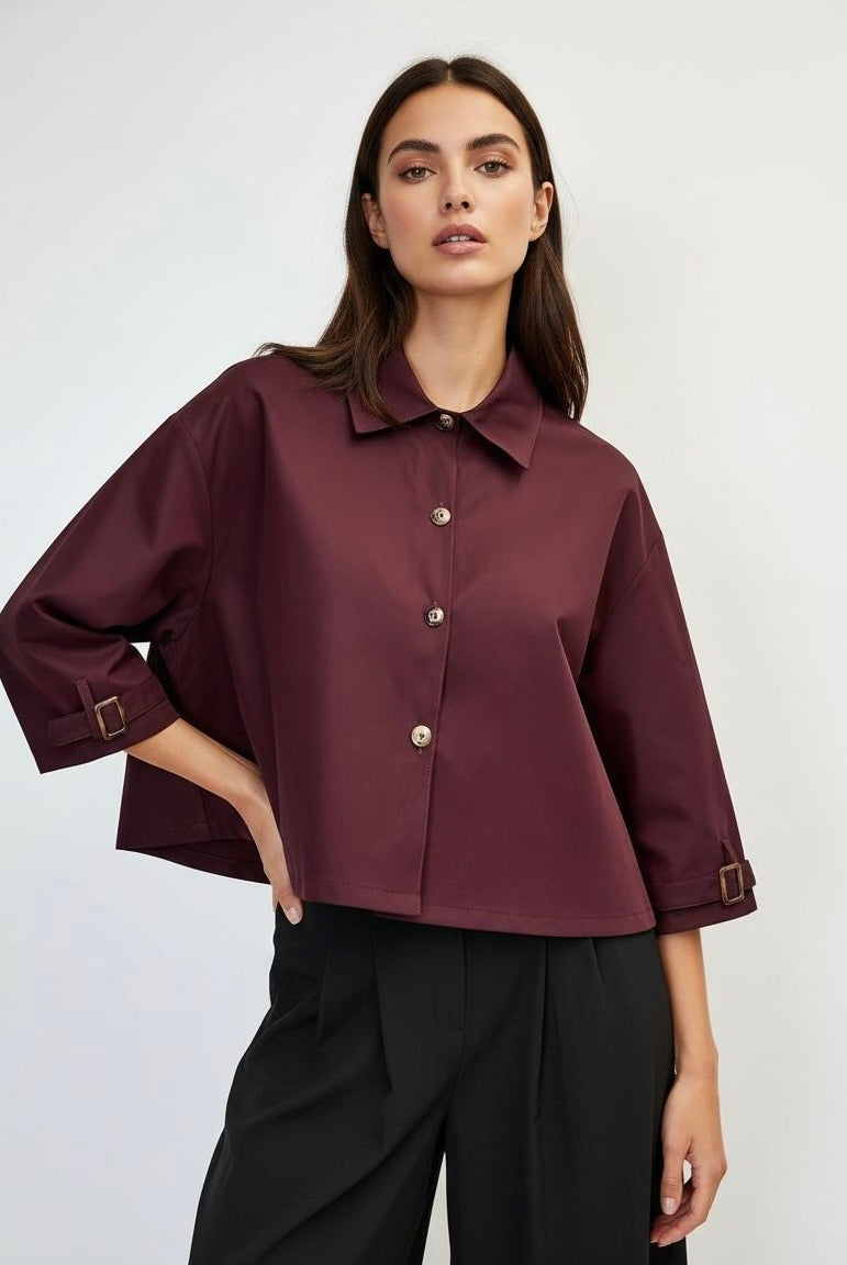 A woman modeling a cropped, long-sleeve button-up jacket in deep burgundy with a collar and buttons, paired with black trousers,