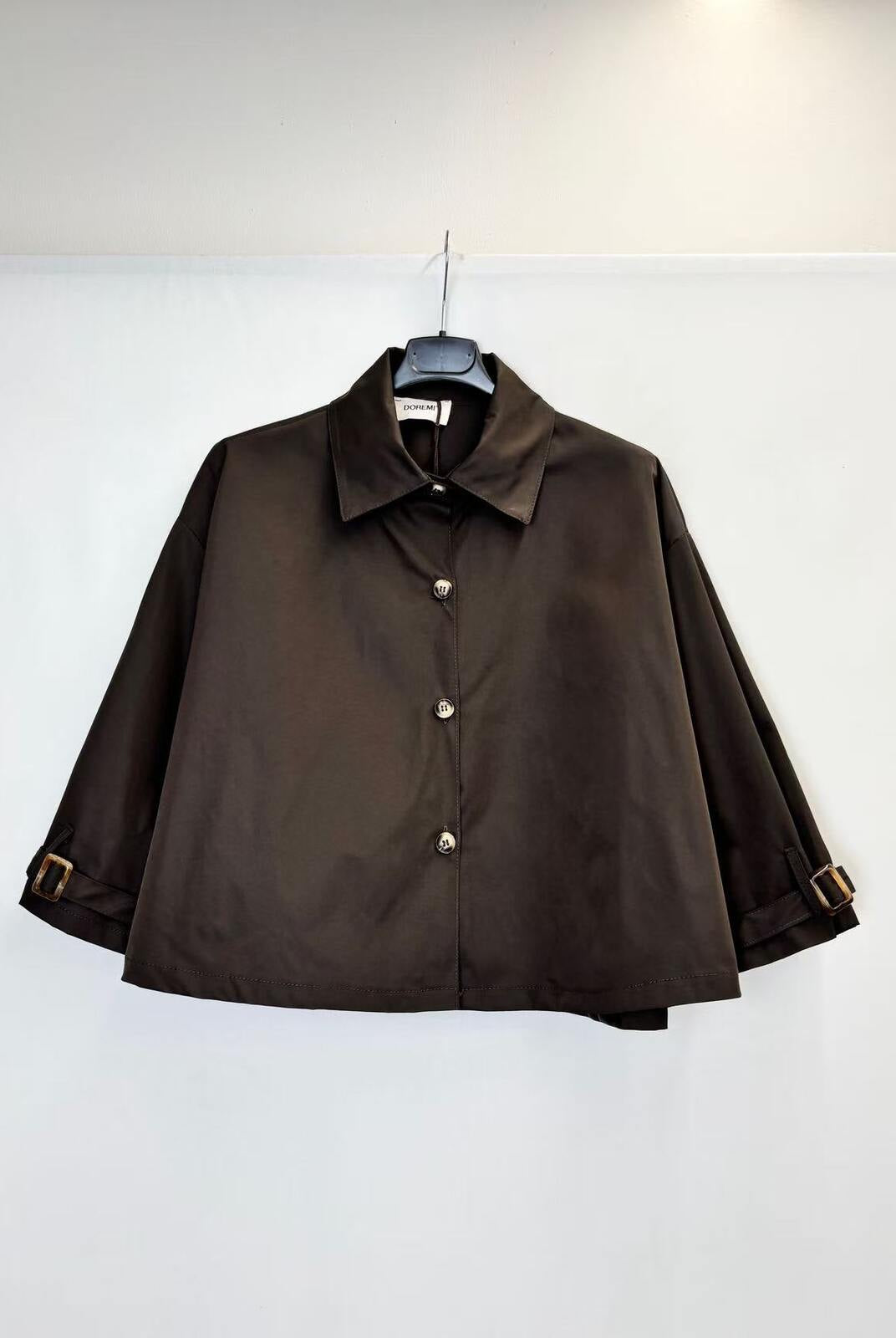 A dark brown trench style jacket with a pointed collar and front buttons, hanging on a black hanger against a plain white wall.