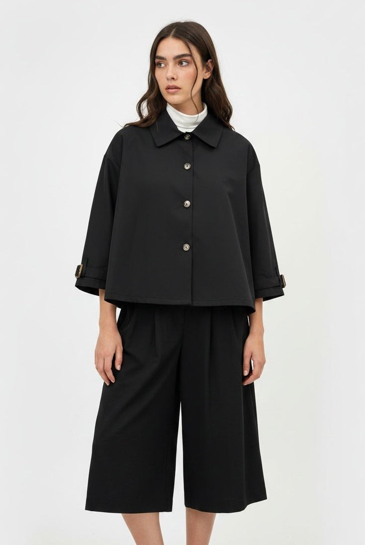A model wearing a black cropped button-up jacket with three-quarter sleeves, paired with matching black wide-leg cropped pants and a white turtleneck underneath