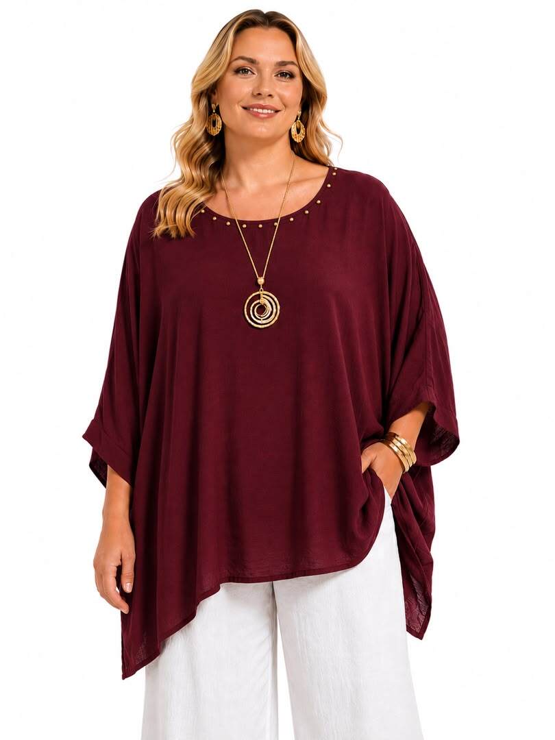 A woman modeling a loose-fitting, scoop-neck top in deep burgundy with small gold stud embellishments around the neckline, paired with white pants and gold jewelry, against a plain white background.