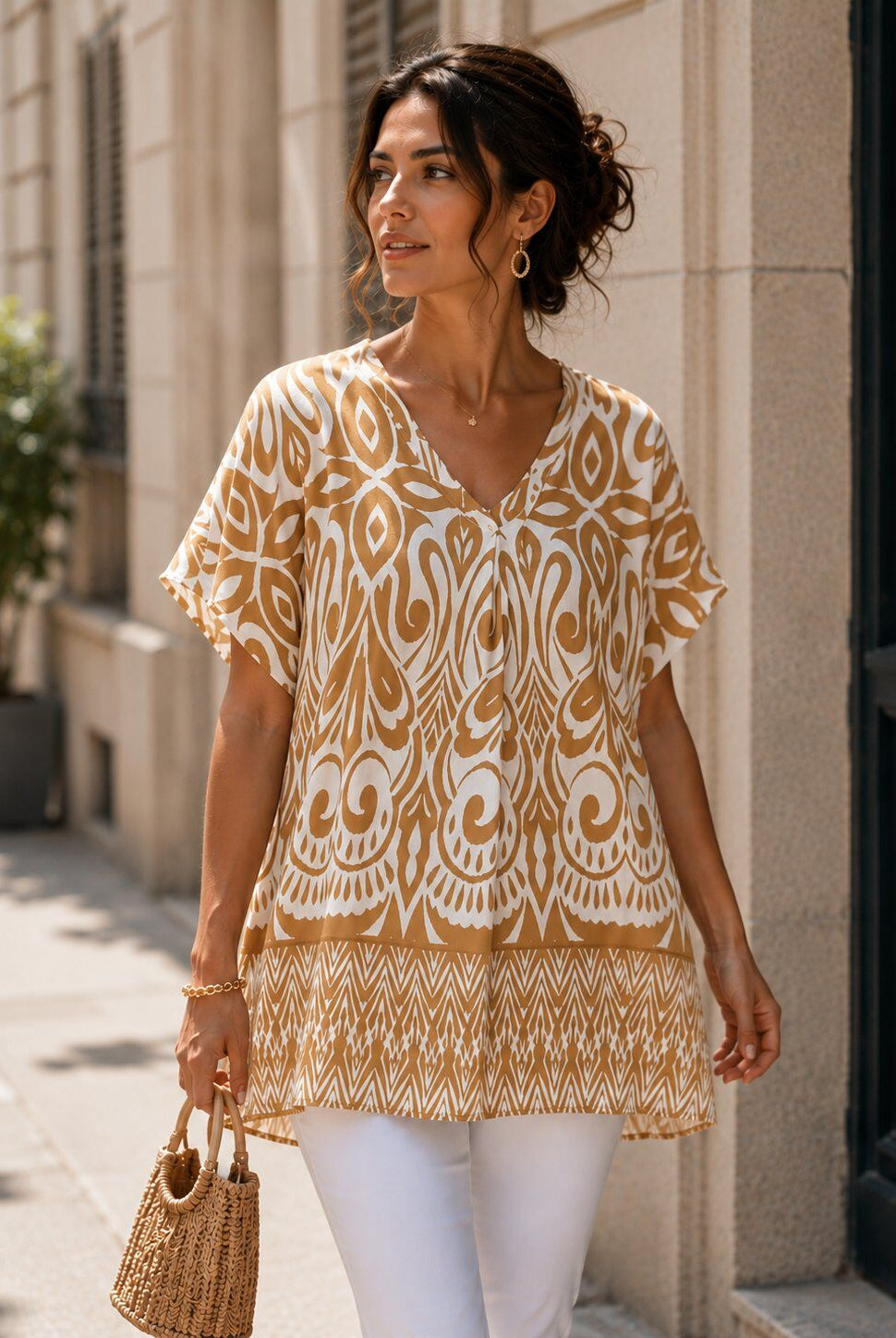 A woman wearing a beige and white patterned short-sleeve tunic top with white pants, walking outdoors on a city sidewalk next to a beige stone building.