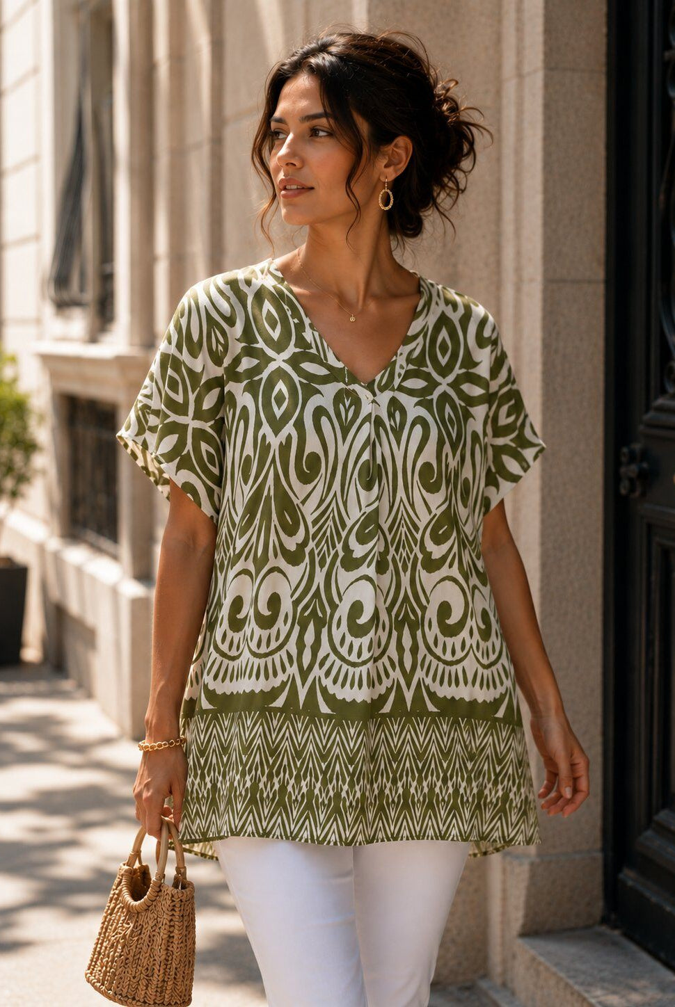 A woman wearing a short-sleeve V-neck tunic top with an olive green and white bohemian-style pattern, paired with white pants and carrying a woven straw handbag, walking outdoors on a city street.