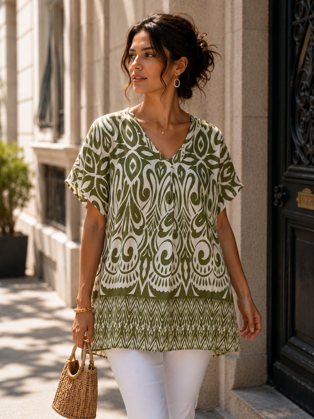 A woman wearing a short-sleeve V-neck tunic top with an olive green and white bohemian-style pattern, paired with white pants and carrying a woven straw handbag, walking outdoors on a city street.