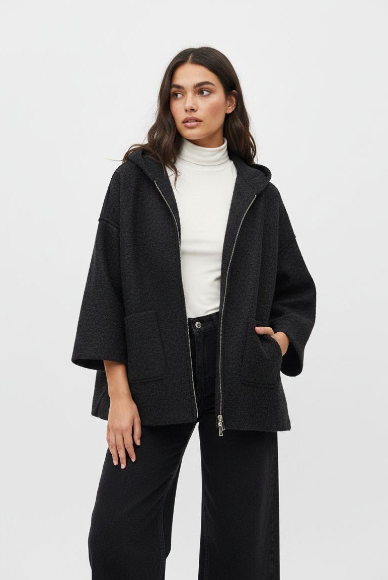 A woman modeling a black textured hooded zip-up jacket with wide sleeves, worn over a white turtleneck and black wide-leg pants