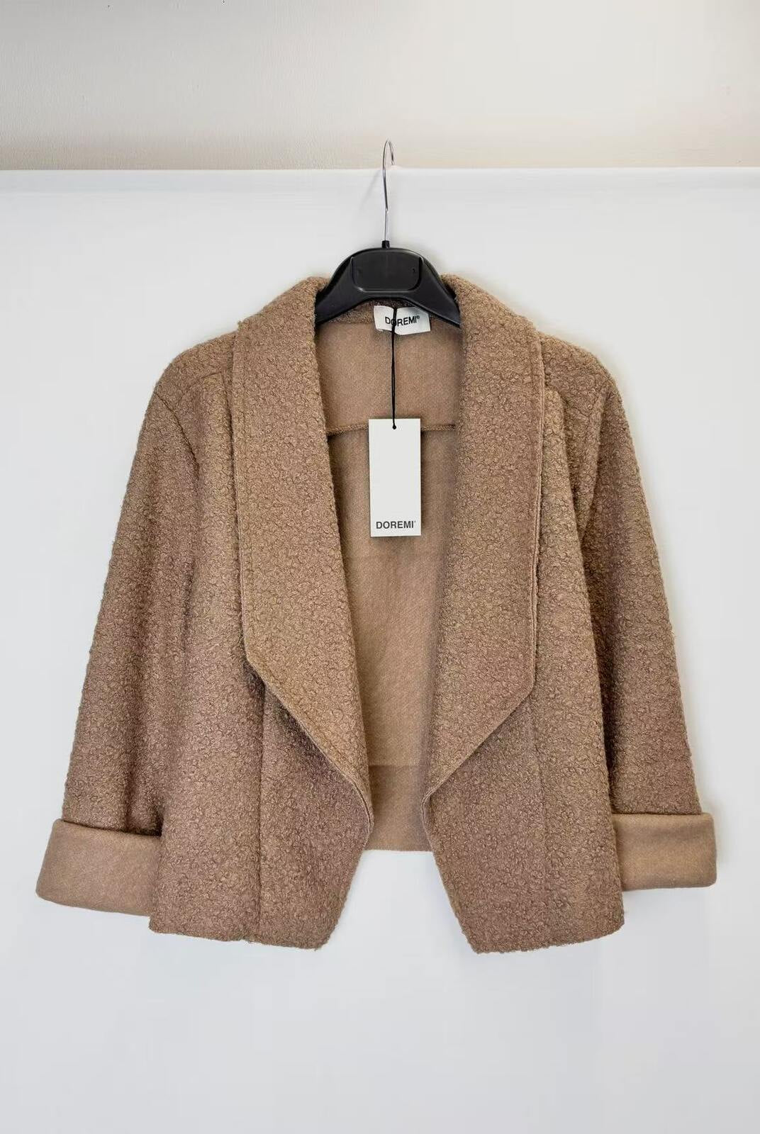 A tan textured bouclé open-front jacket with wide lapels and rolled cuffs, hanging on a black hanger