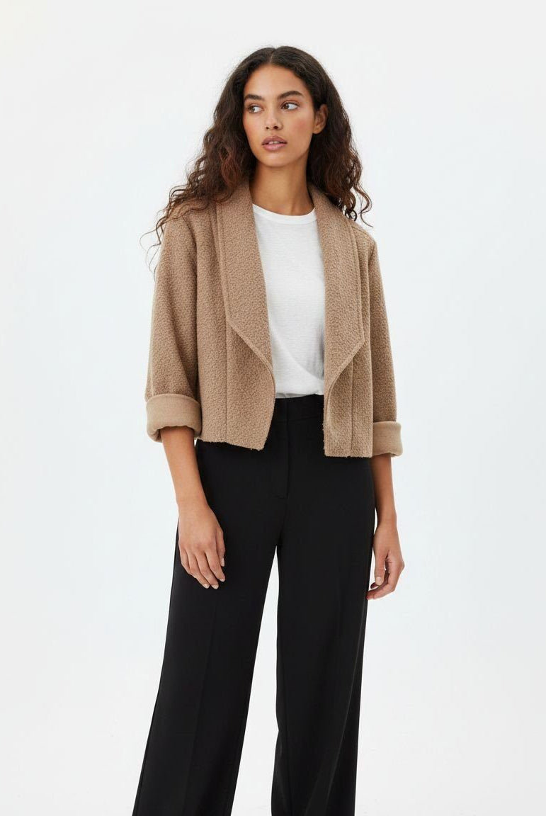 A woman modeling a cropped, open-front beige bouclé jacket with rolled cuffs over a white top and black wide-leg trousers, photographed against a clean white studio background.