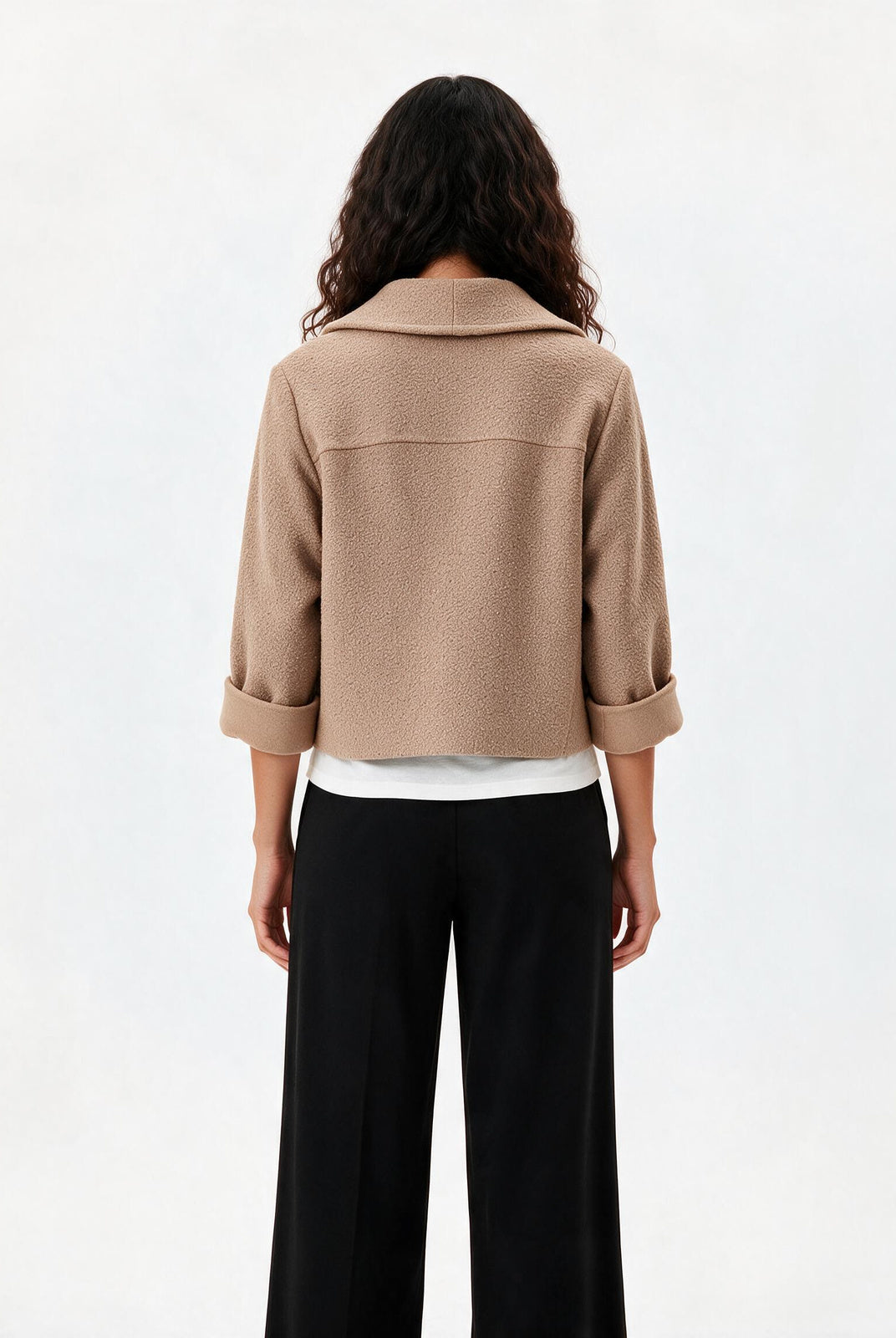 A model shown from the back wearing a cropped beige jacket with rolled three-quarter sleeves, black wide-leg trousers, and a white undershirt, against a plain white studio background.