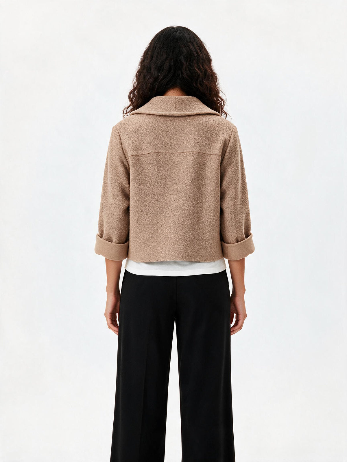 A model shown from the back wearing a cropped beige jacket with rolled three-quarter sleeves, black wide-leg trousers, and a white undershirt, against a plain white studio background.