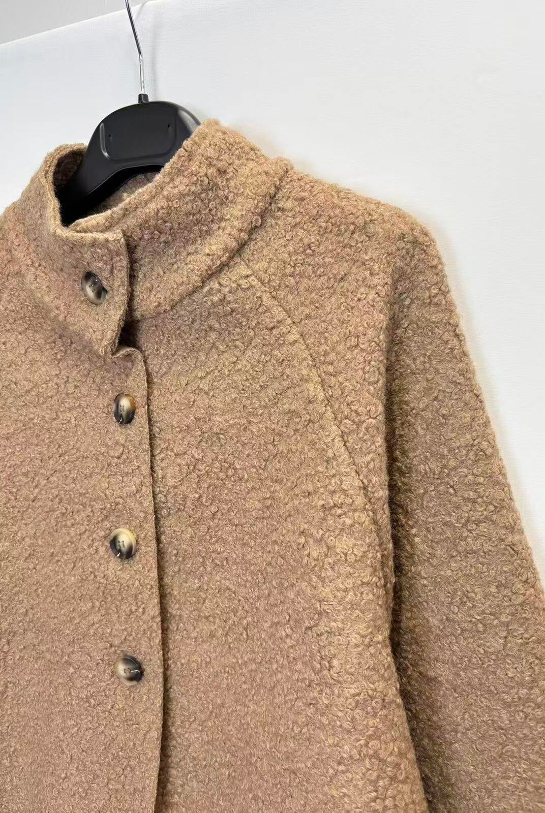 A close-up of a textured, bouclé-like brown jacket hanging on a black hanger against a plain white wall.