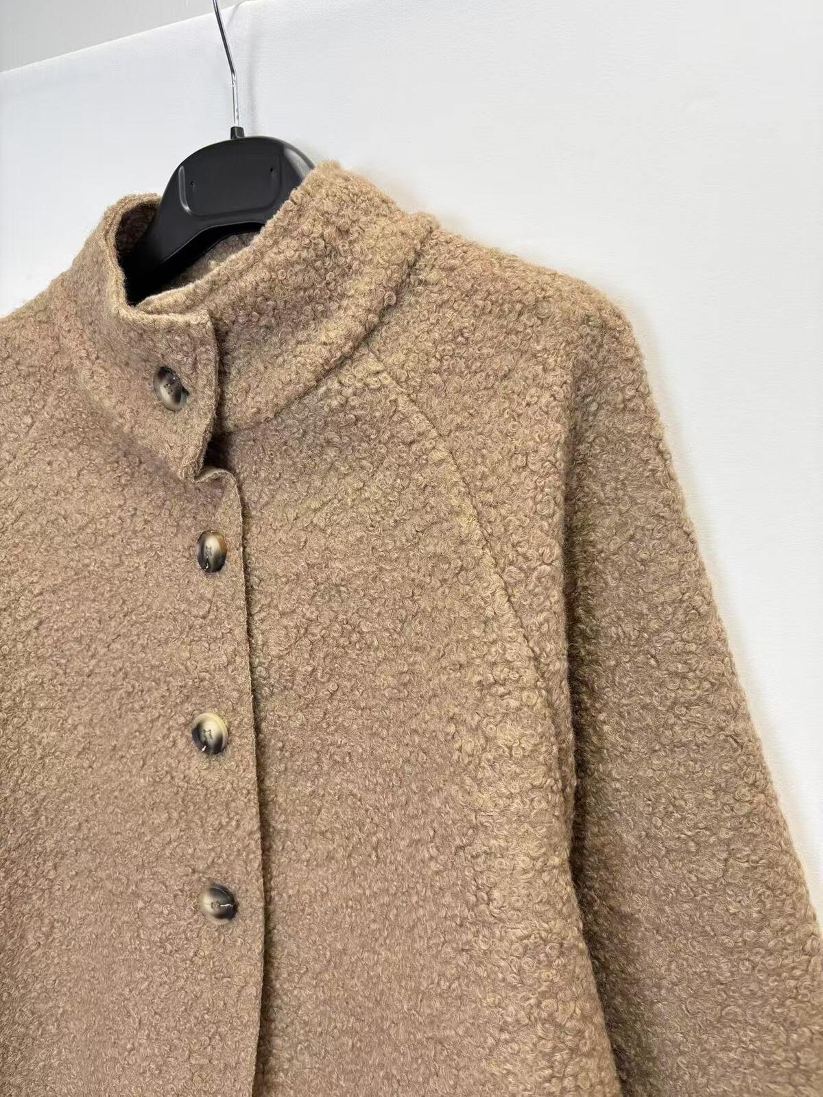 A close-up of a textured, bouclé-like brown jacket hanging on a black hanger against a plain white wall.