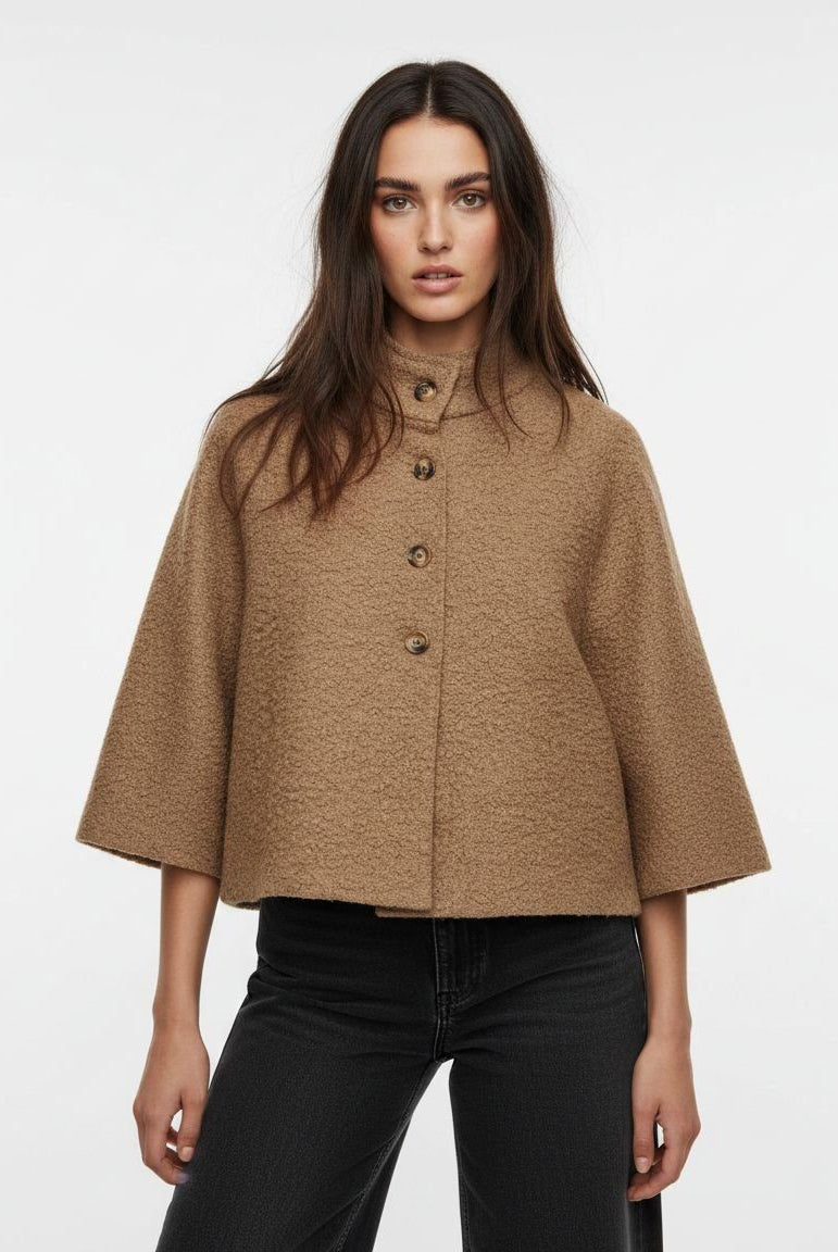 A woman modeling a cropped, high-neck, short-sleeved cape jacket in a textured camel brown fabric, paired with black jeans, against a plain white studio background.