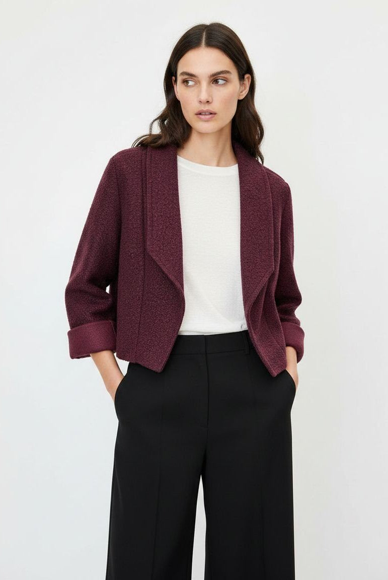 A woman modeling a textured, open-front cropped jacket in deep burgundy, worn over a white top and black wide-leg trousers, against a plain light gray studio background.