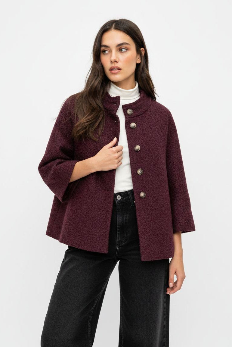 A woman modeling a short, textured burgundy jacket with a high stand collar and decorative silver-tone buttons, worn over a white turtleneck and black wide-leg jeans, against a plain white studio background.