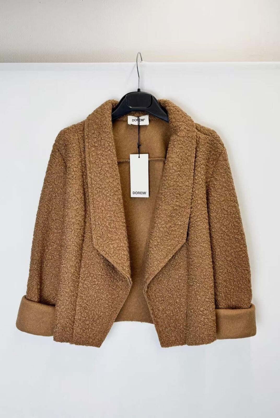 A camel brown bouclé cropped jacket with wide lapels and rolled cuffs, hanging on a black hanger