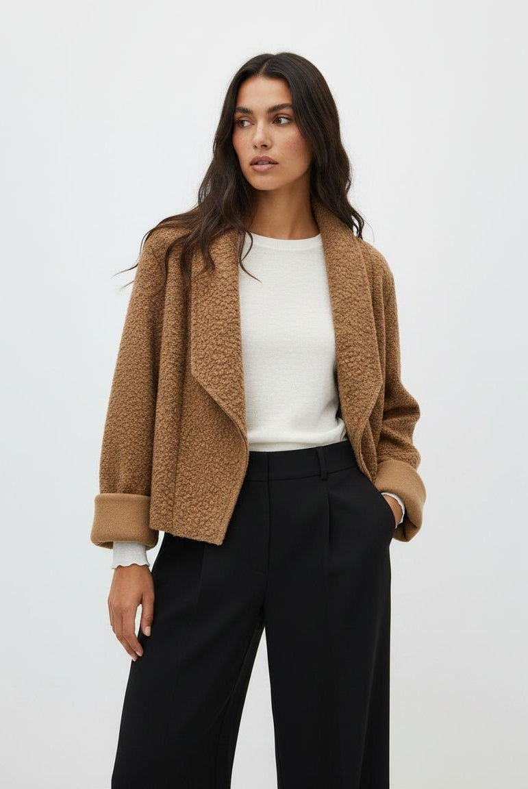 A woman modeling a cropped, open-front teddy fleece jacket in camel brown, worn over a white ribbed top and black wide-leg trousers, against a plain light gray studio background.