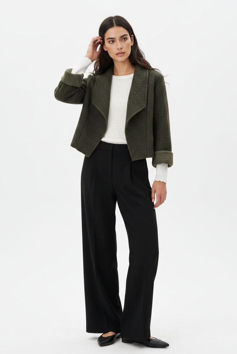 A woman modeling a cropped olive green textured jacket with rolled cuffs, worn over a white long-sleeve top and black wide-leg trousers, against a plain white studio background.