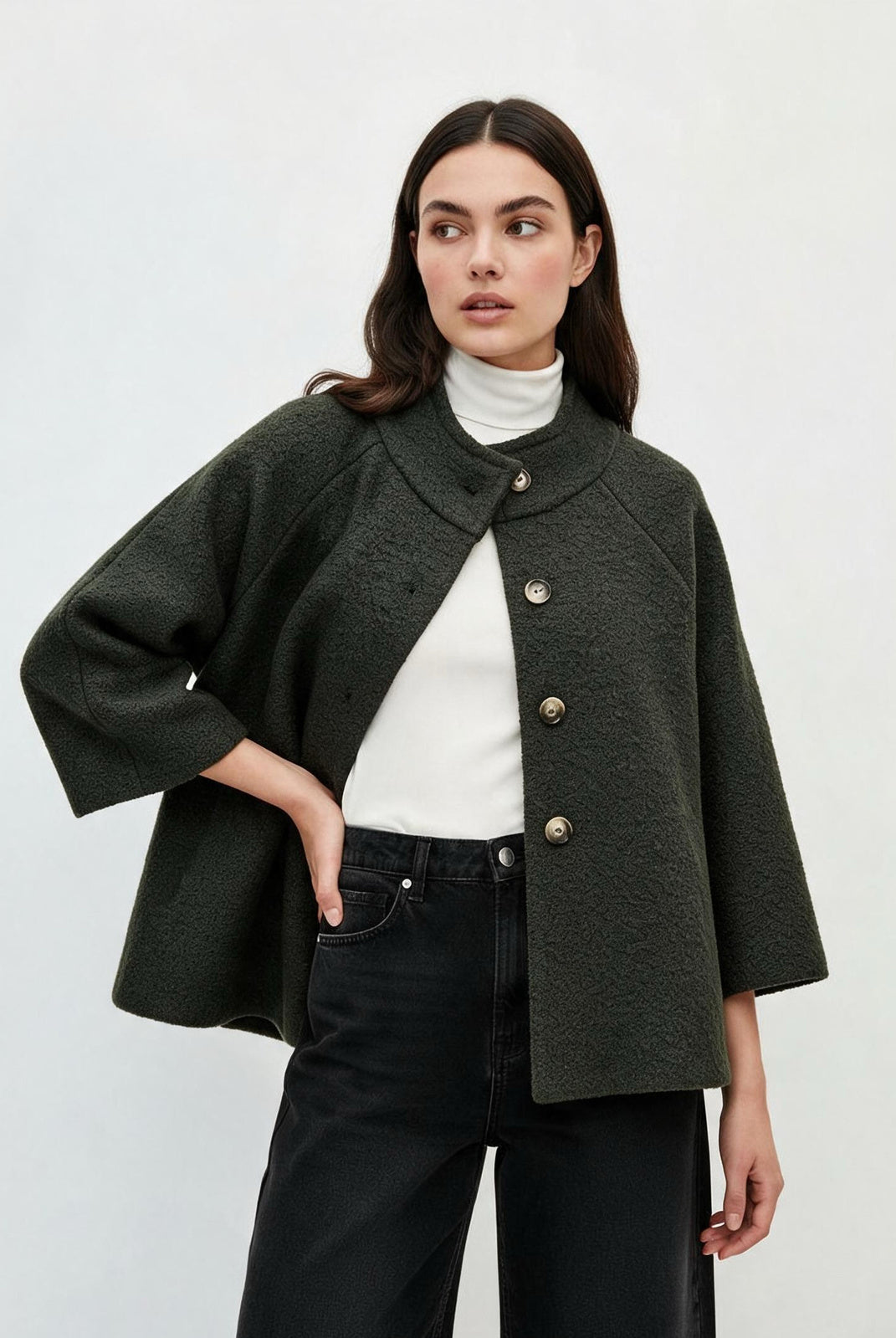 A woman modeling a dark olive green textured cropped jacket with wide three-quarter sleeves and gold-tone buttons, worn over a white turtleneck and black jeans against a light gray studio background.
