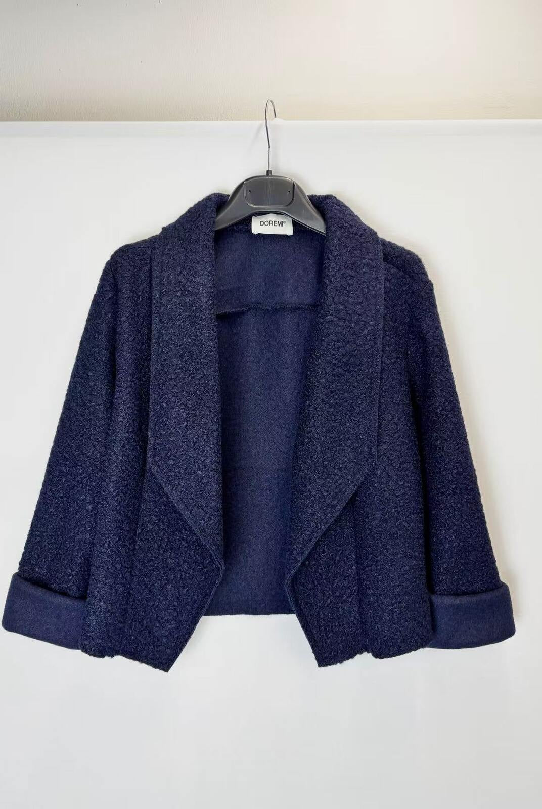 A navy blue textured bouclé jacket with wide lapels and rolled cuffs, hanging on a black hanger against a plain white wall.