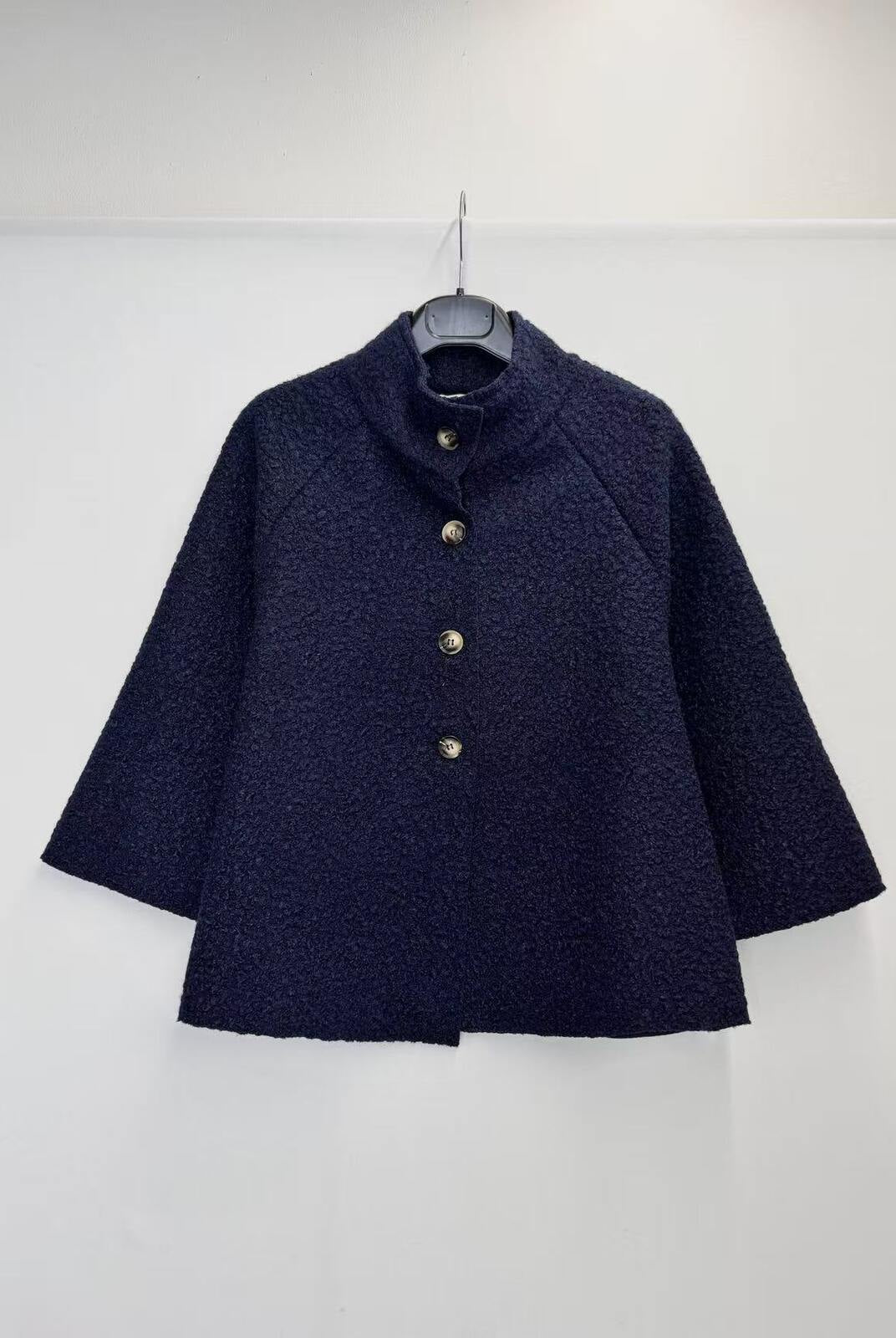 A dark navy blue textured jacket with a high mock neck and gold buttons, hanging on a black hanger against a plain white wall.