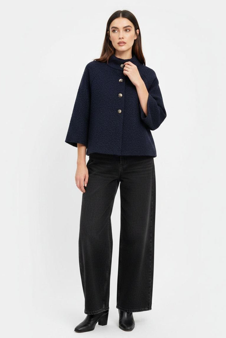 A model wearing a dark navy blue cropped jacket with three-quarter sleeves and gold buttons, paired with black wide-leg trousers and black boots, against a plain white studio background.