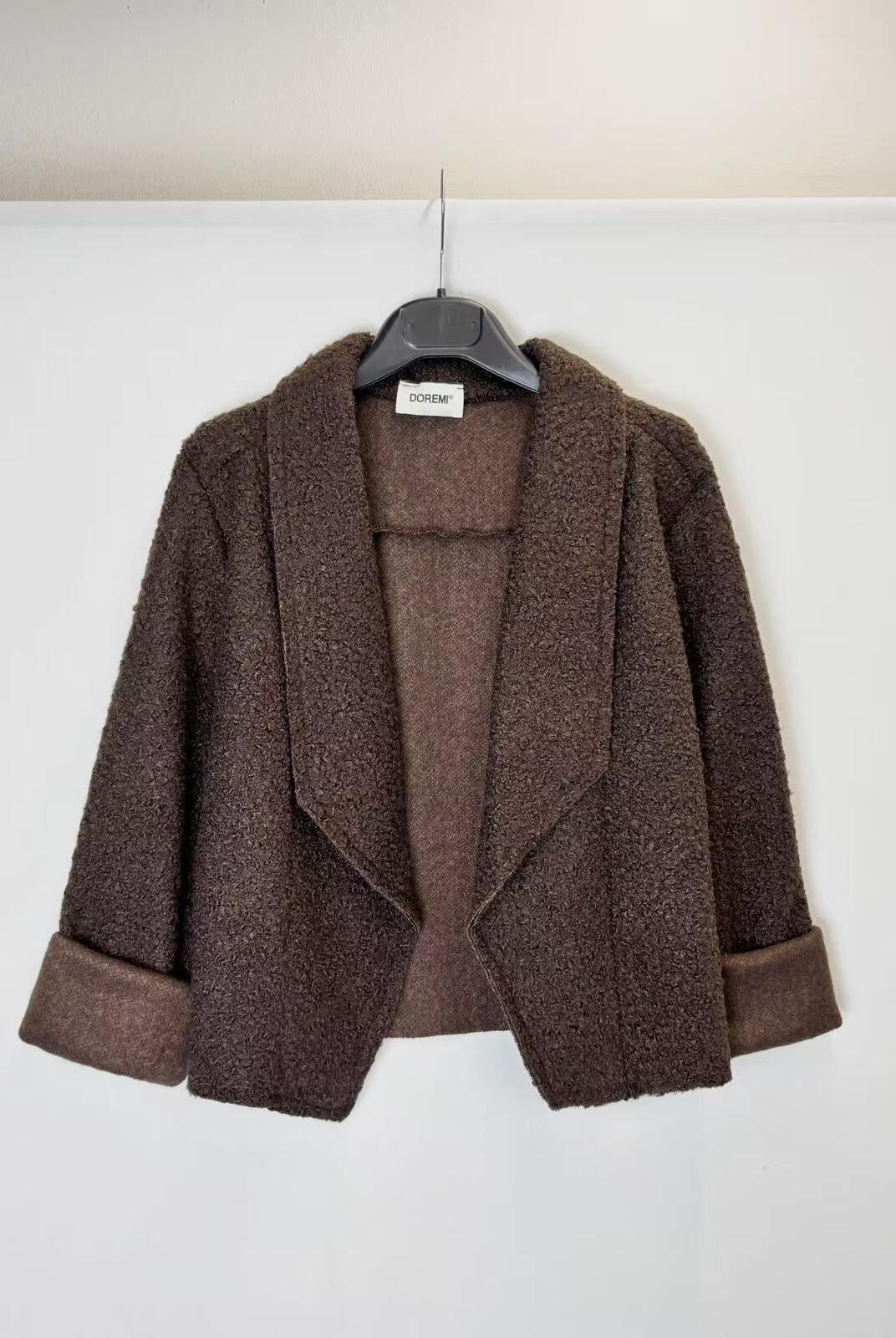 A brown textured open-front jacket with wide lapels and rolled cuffs, hanging on a black hanger against a plain white wall.
