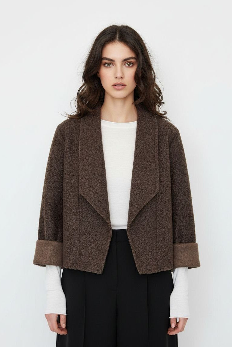 A woman modeling a cropped brown textured jacket with wide lapels and turned-back cuffs, worn over a white top and black trousers against a plain light gray background.