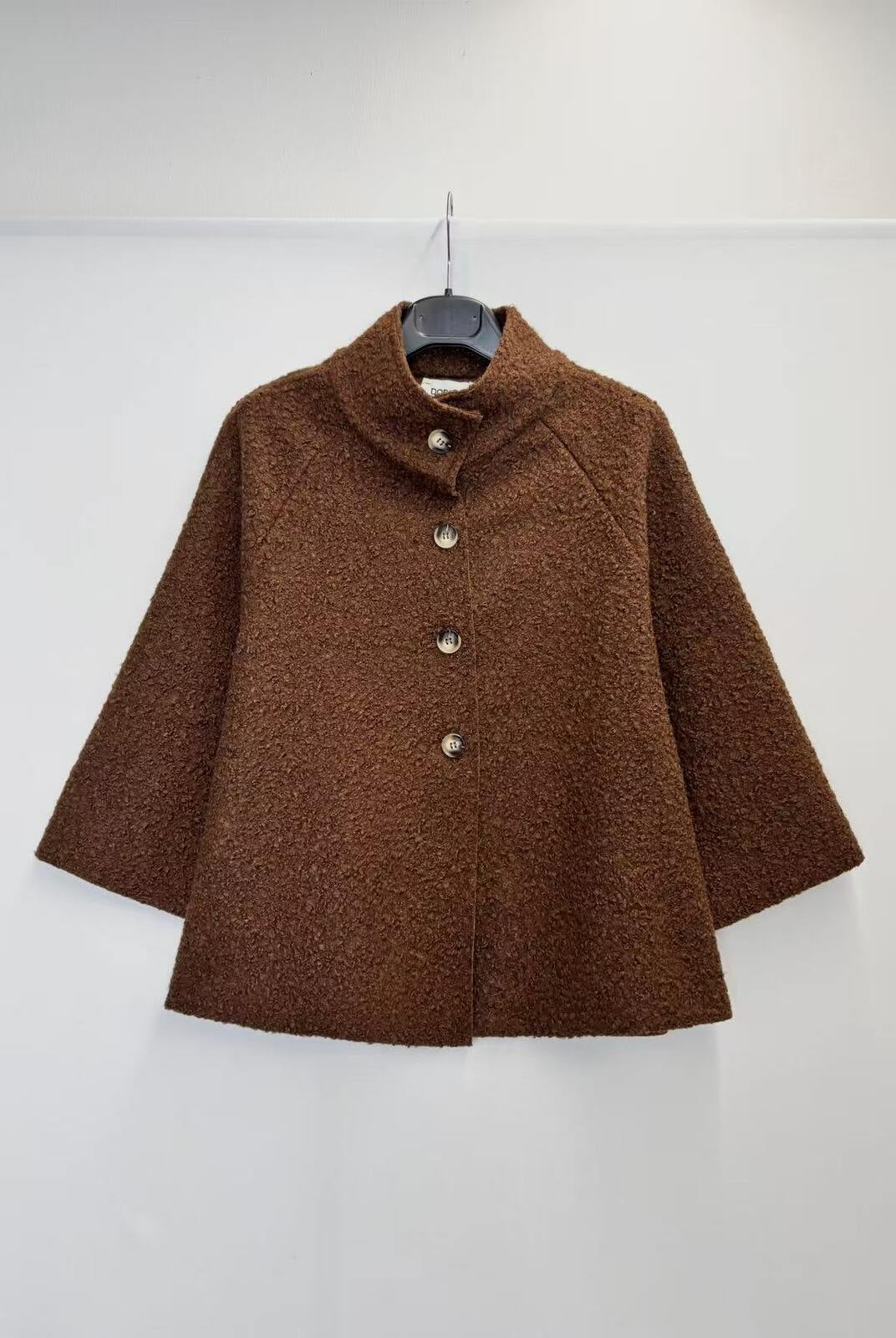 A brown textured bouclé jacket with a mock neck and three front buttons, hanging on a black hanger against a plain white wall.