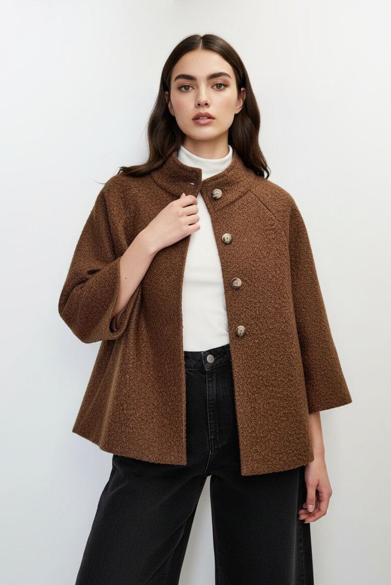 A woman modeling a brown textured bouclé jacket with a mock neck and decorative buttons, worn over a white turtleneck and black wide-leg jeans, against a plain white studio background.