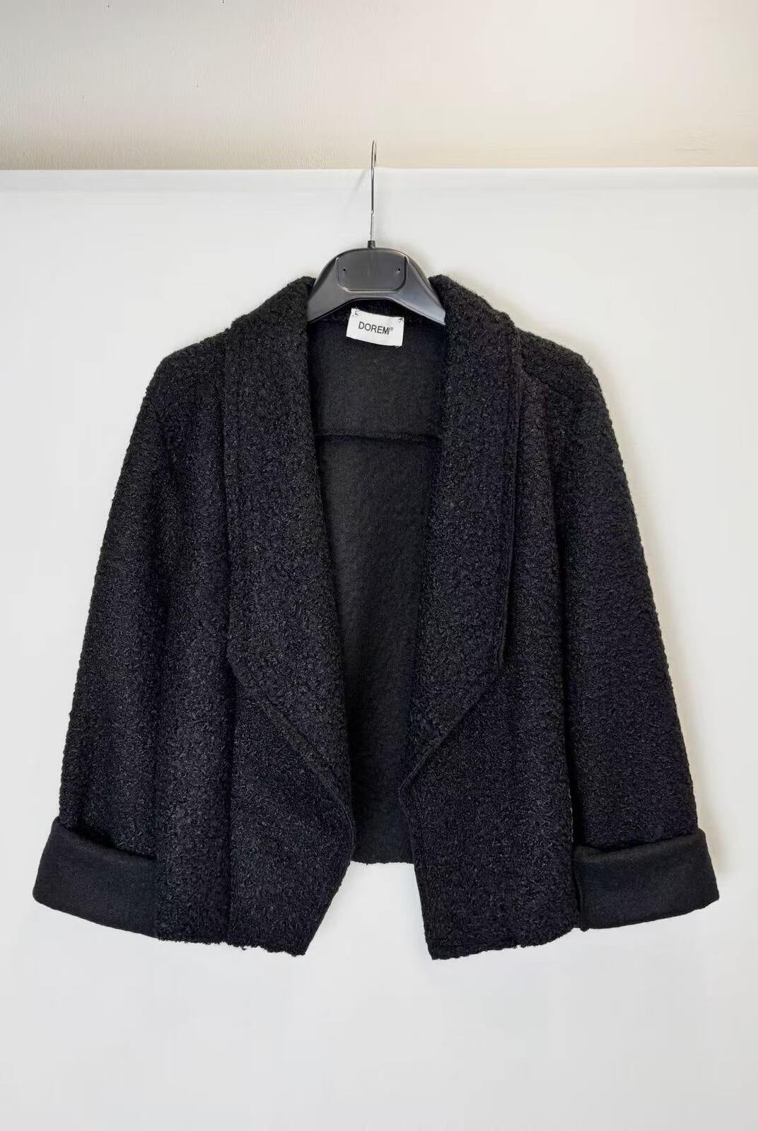 A black textured bouclé jacket with wide lapels and rolled cuffs, hanging on a black hanger against a plain white wall.