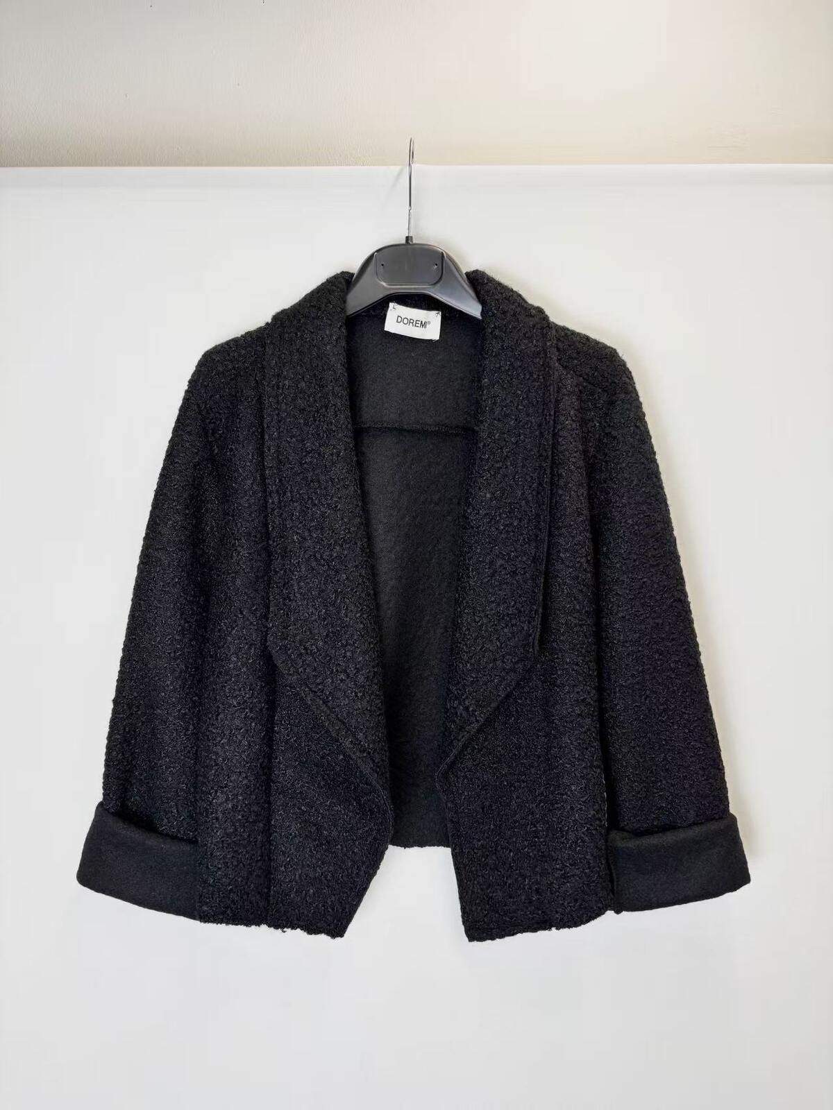 A black textured bouclé jacket with wide lapels and rolled cuffs, hanging on a black hanger against a plain white wall.