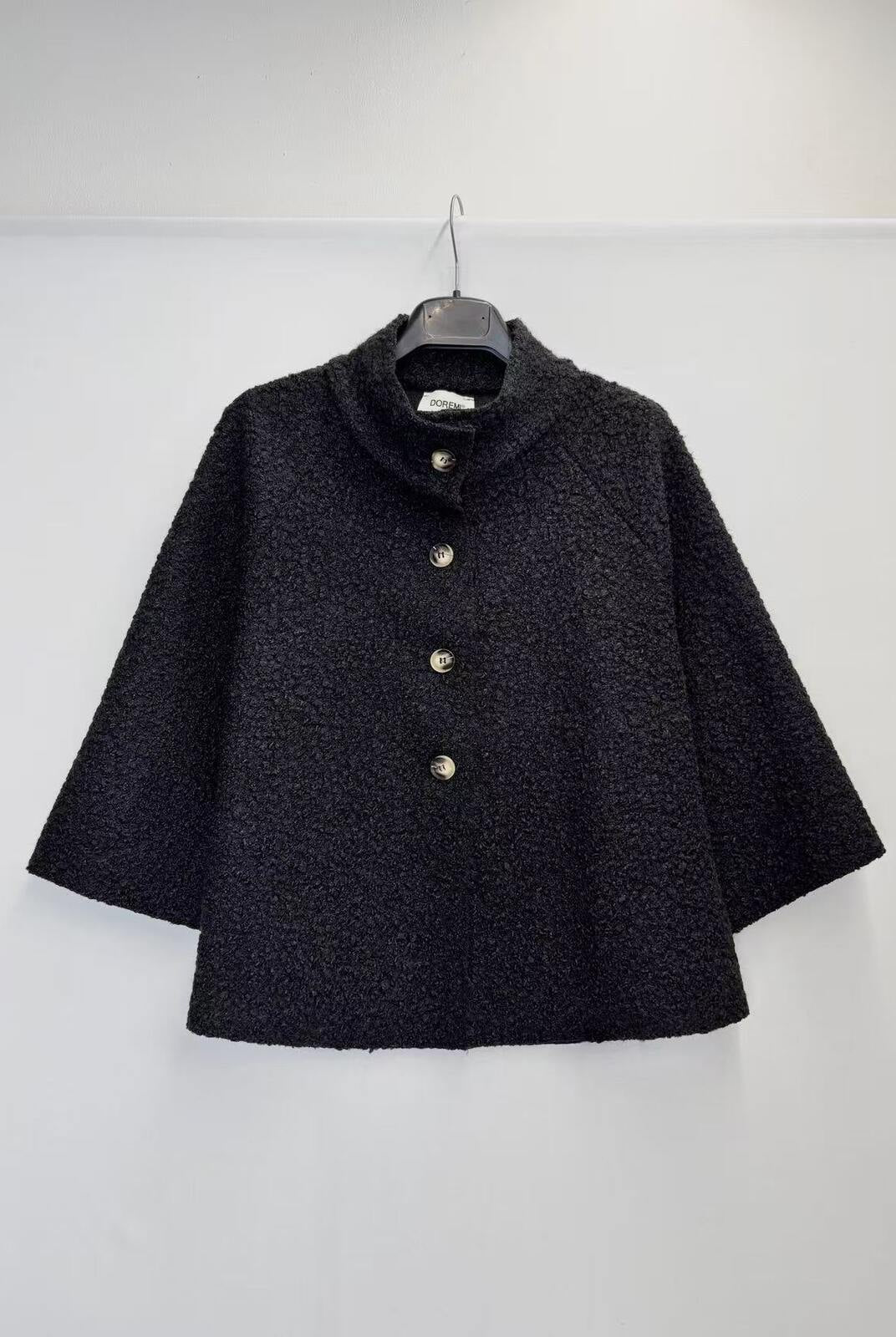 A black textured bouclé cape jacket with a high stand collar and three gold-tone buttons, hanging on a black hanger against a plain white wall.