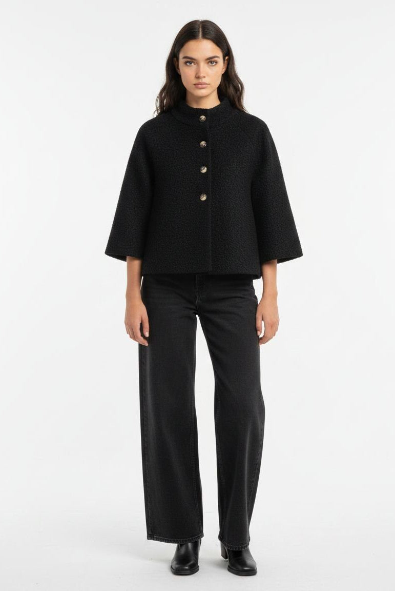 A model stands against a plain white studio background wearing a black cropped cape jacket with three-quarter sleeves and gold-tone buttons, paired with black wide-leg trousers and black shoes.