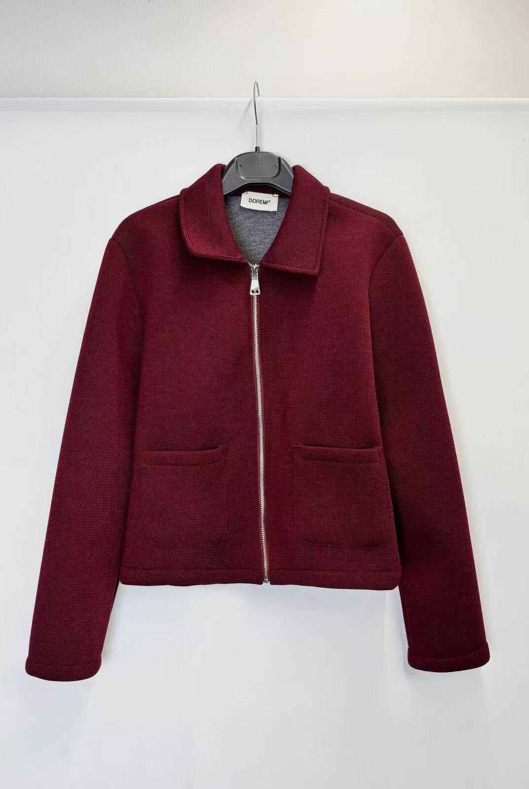 A maroon zip-up jacket with a stand collar and two front patch pockets, hanging on a black hanger against a plain white wall.