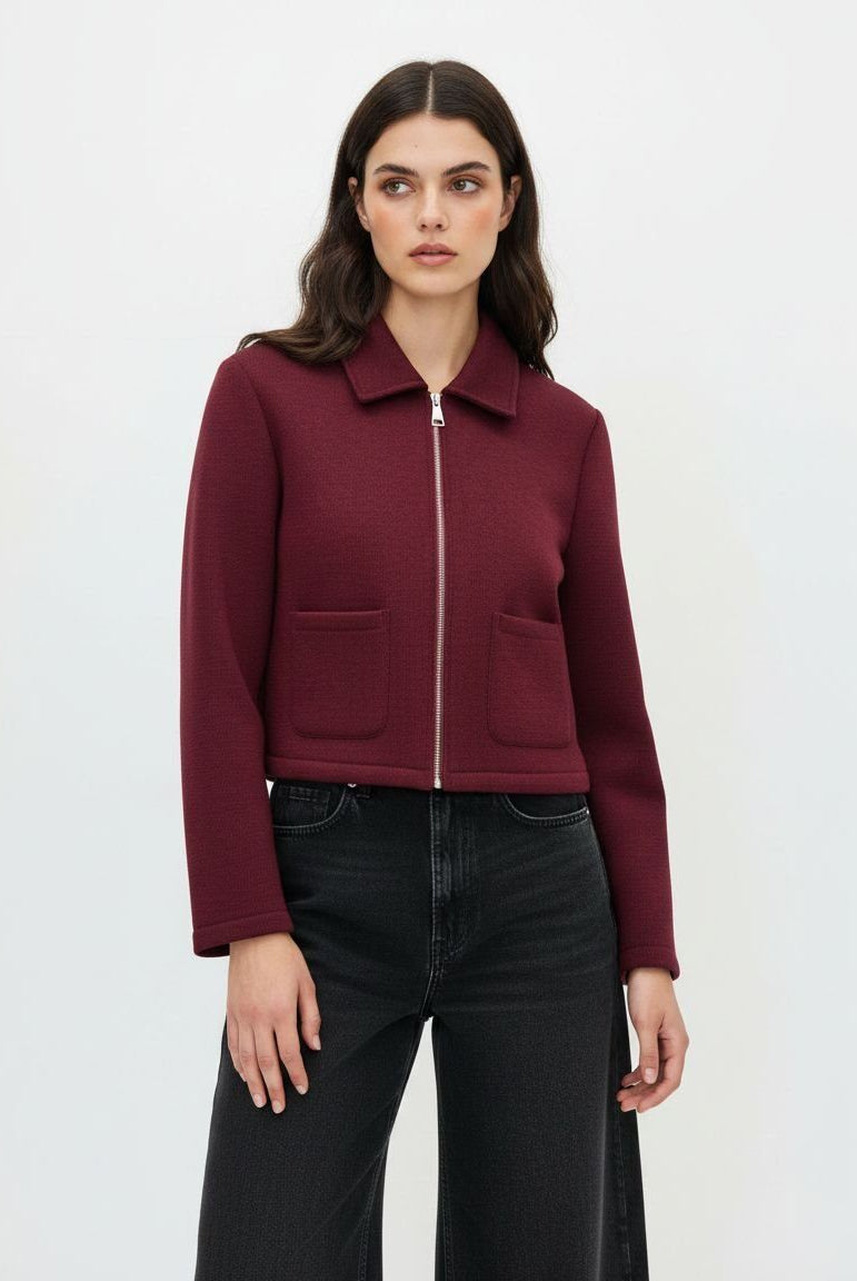 A woman modeling a cropped, long-sleeve zip-up jacket in deep burgundy with two front patch pockets, paired with black wide-leg jeans, against a plain white studio background.