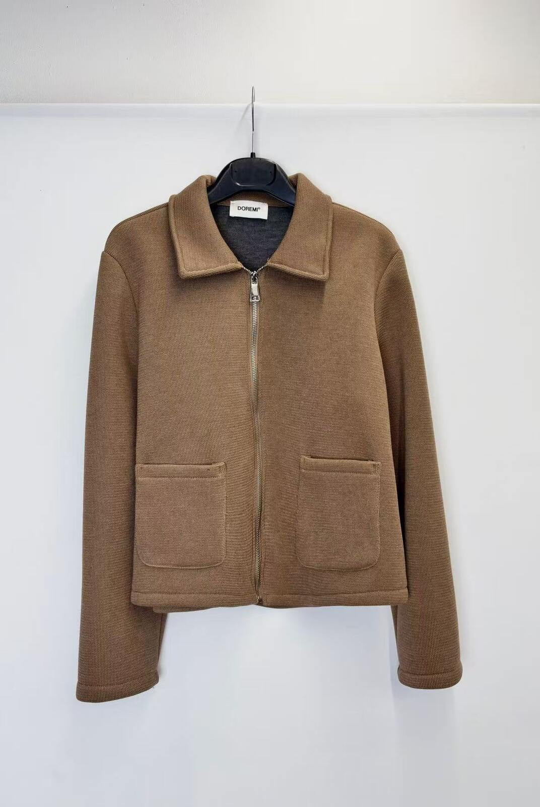 A brown zip-up jacket with a collar and two front patch pockets, hanging on a black hanger against a plain white wall.