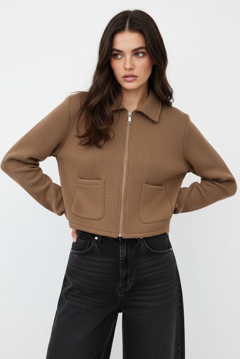 A woman modeling a cropped, long-sleeve zip-front jacket in a warm camel brown color, paired with black jeans, against a plain light gray studio background.