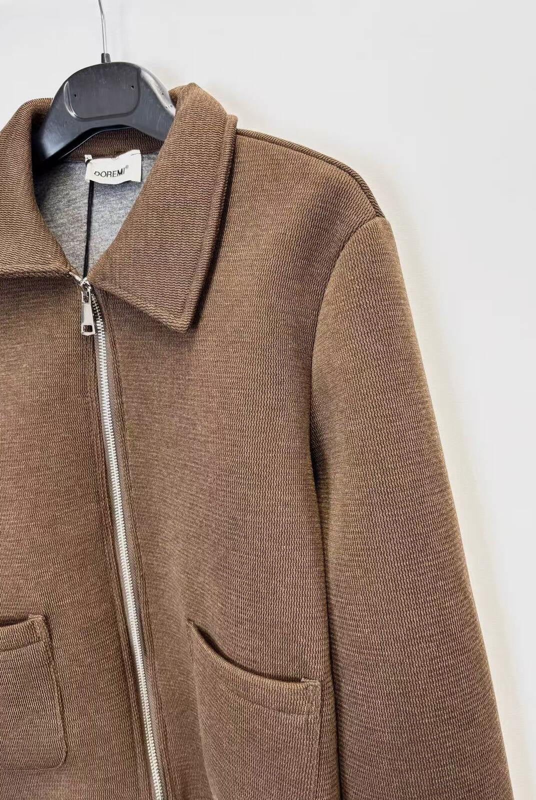 A brown ribbed knit zip-up jacket with a collar and front pockets, hanging on a black hanger against a plain white wall.
