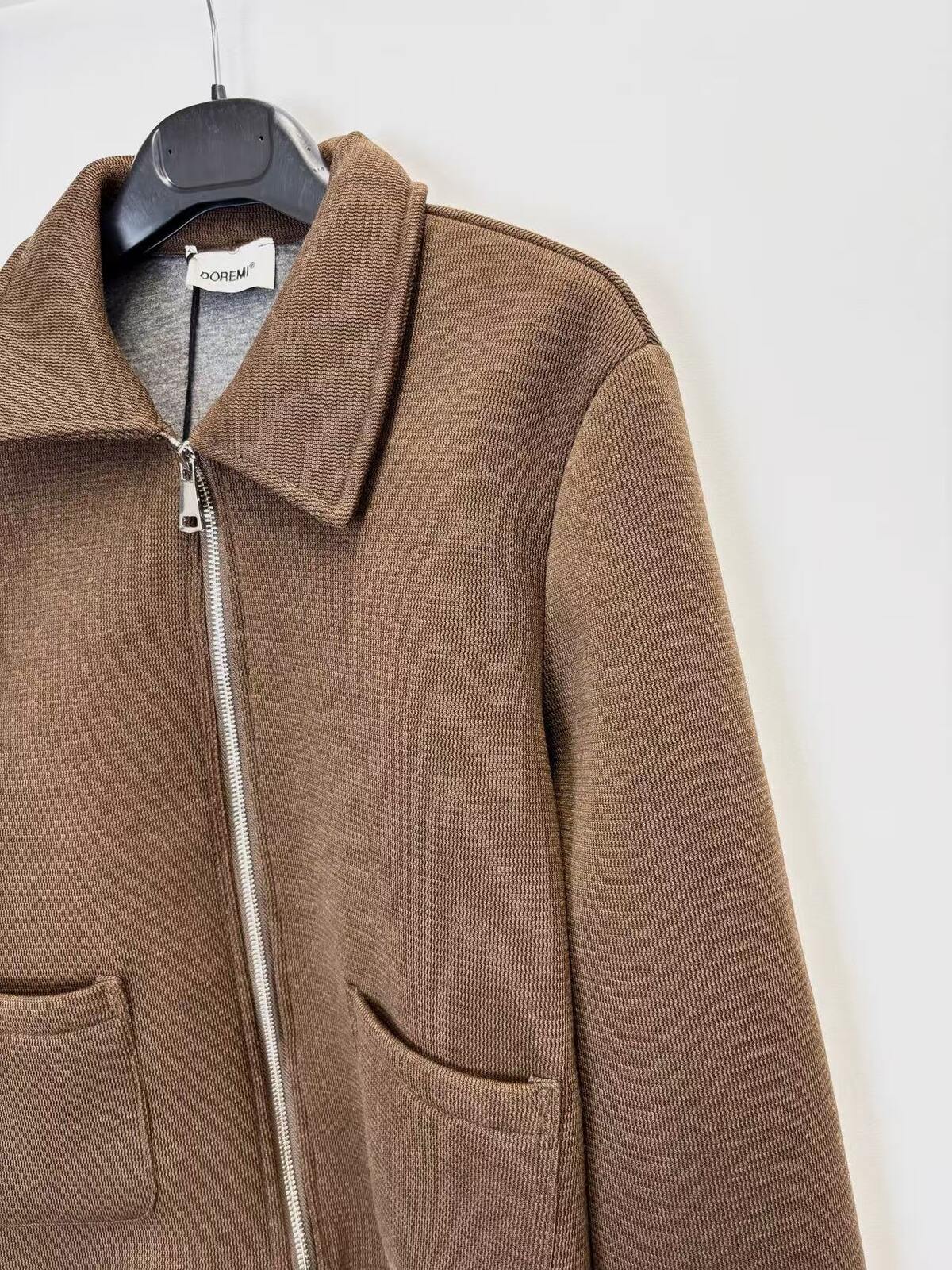 A brown ribbed knit zip-up jacket with a collar and front pockets, hanging on a black hanger against a plain white wall.