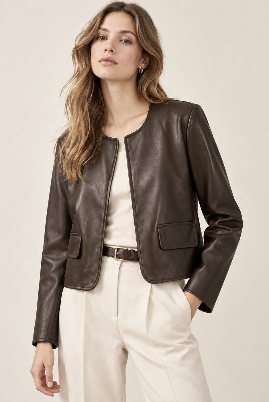 A woman modeling a cropped dark brown faux-leather jacket over a cream top and cream trousers, photographed against a plain light beige studio background.