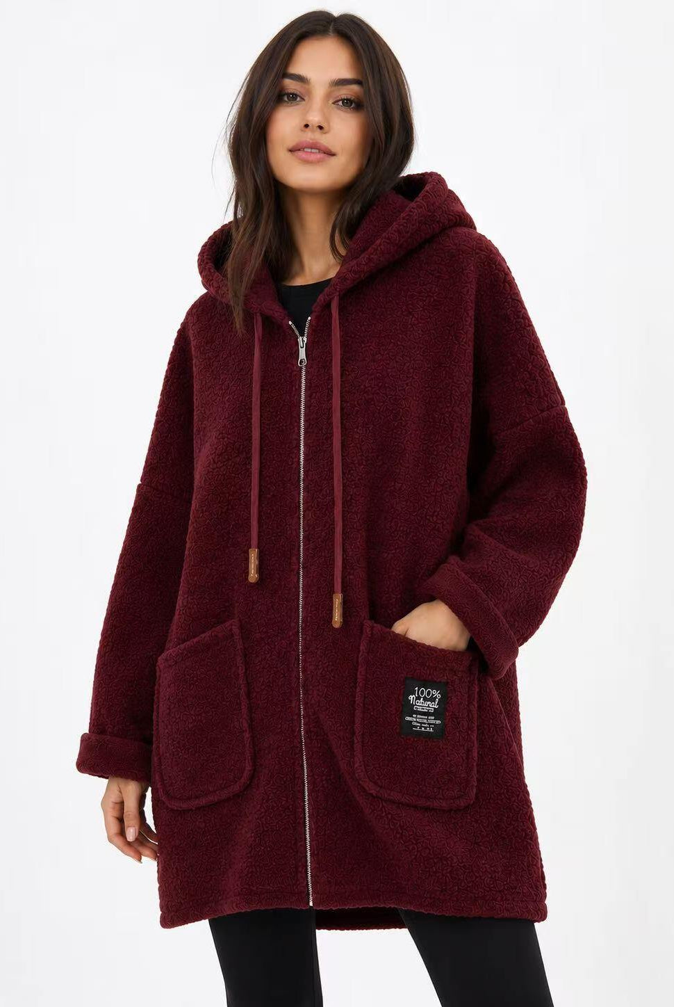A woman wearing a plush, sherpa-textured hooded zip-up coat in deep burgundy. The coat features a drawstring hood, front pockets, and a small black brand patch on the lower right pocket.