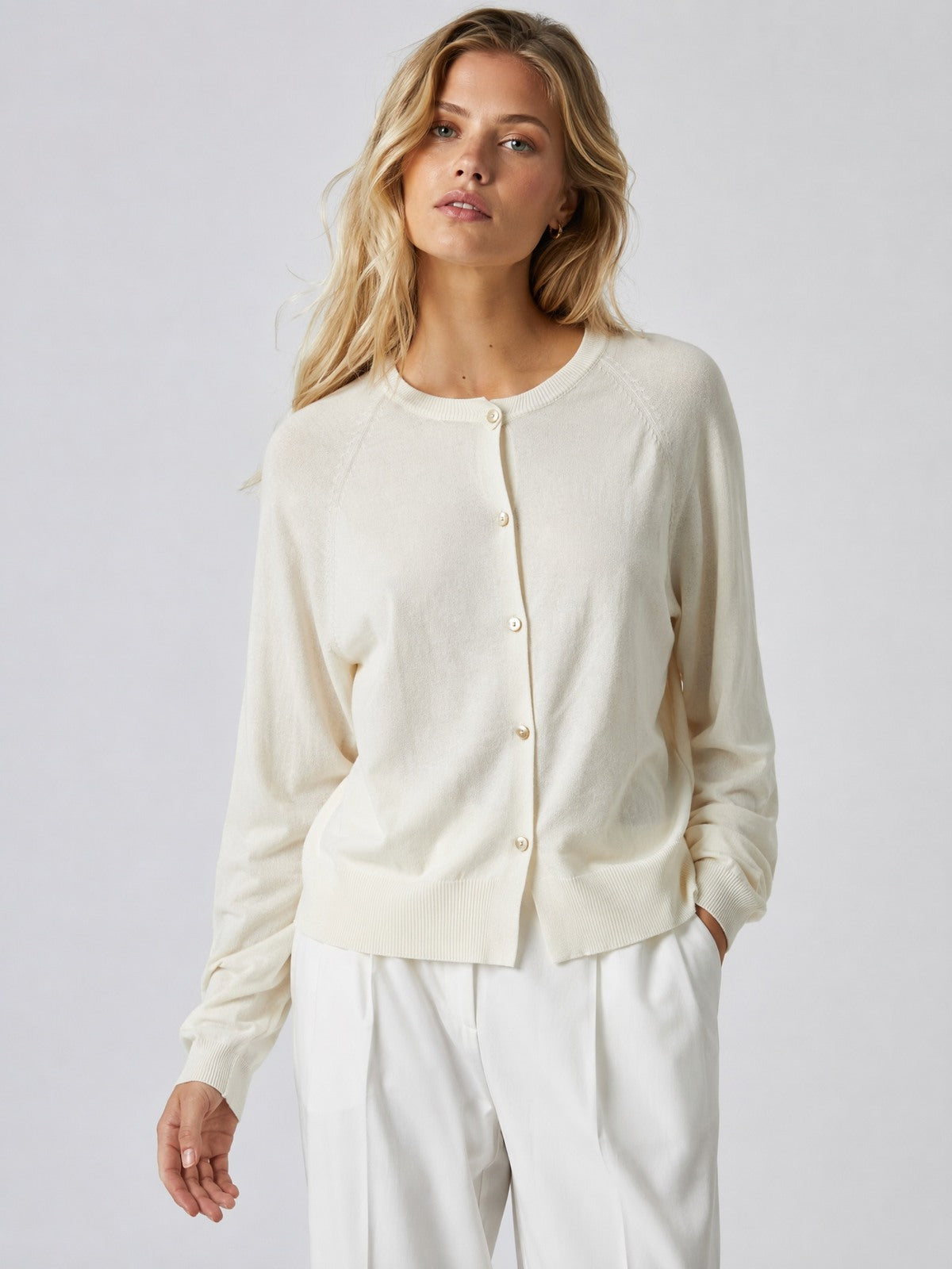 A woman modeling a cream-colored, long-sleeve button-up cardigan sweater paired with white trousers