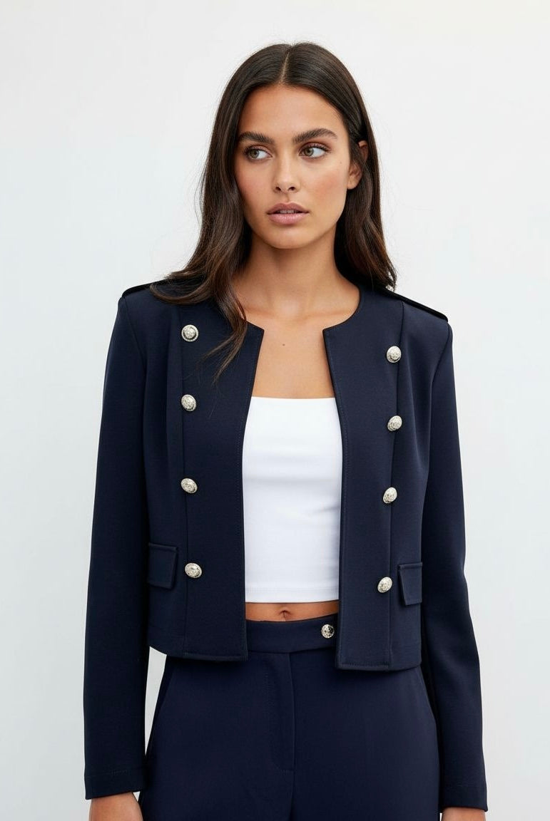 A woman modeling a cropped navy blue double-breasted blazer with silver-tone buttons, worn over a white crop top and matching navy trousers, against a light gray studio background.