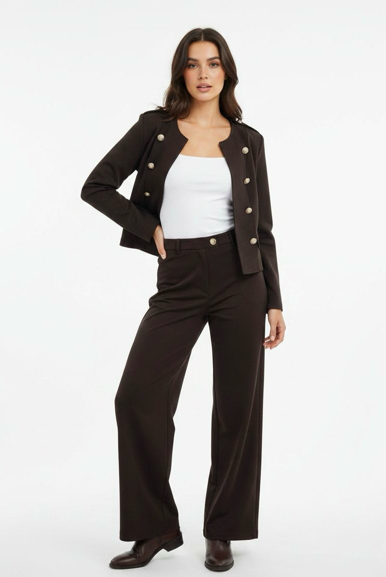A woman modeling a dark brown cropped double-breasted jacket with buttons, matching dark brown wide-leg trousers, a white tank top, and dark brown boots, set against a plain white studio background.