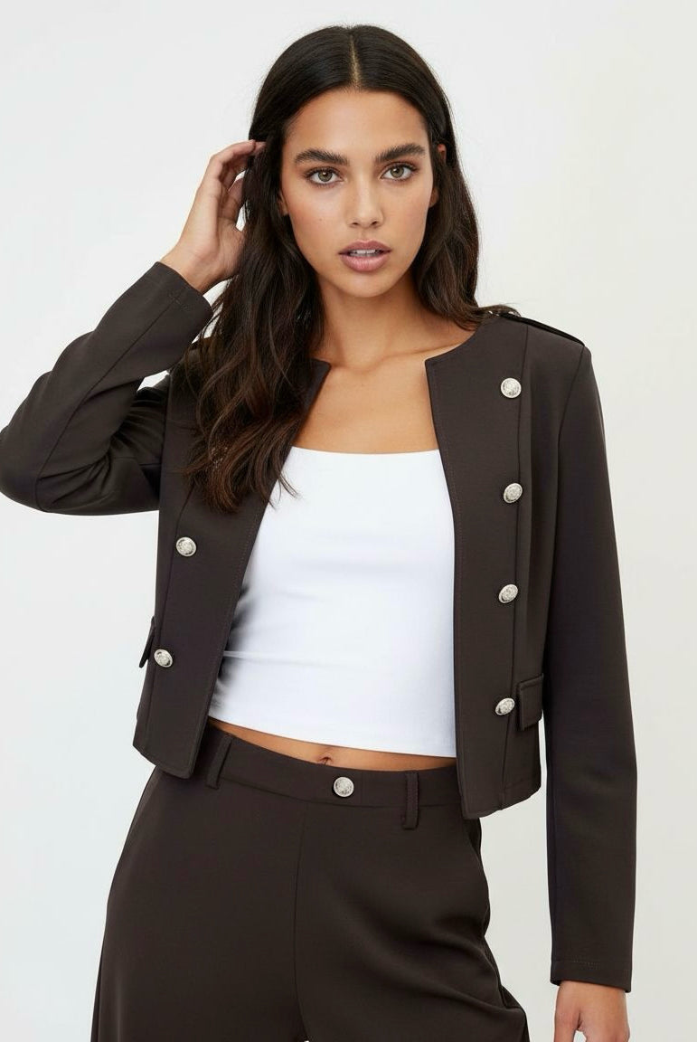 A woman modeling a dark brown cropped double-breasted blazer with silver buttons, paired with matching dark brown trousers and a white crop top, against a plain light gray background.