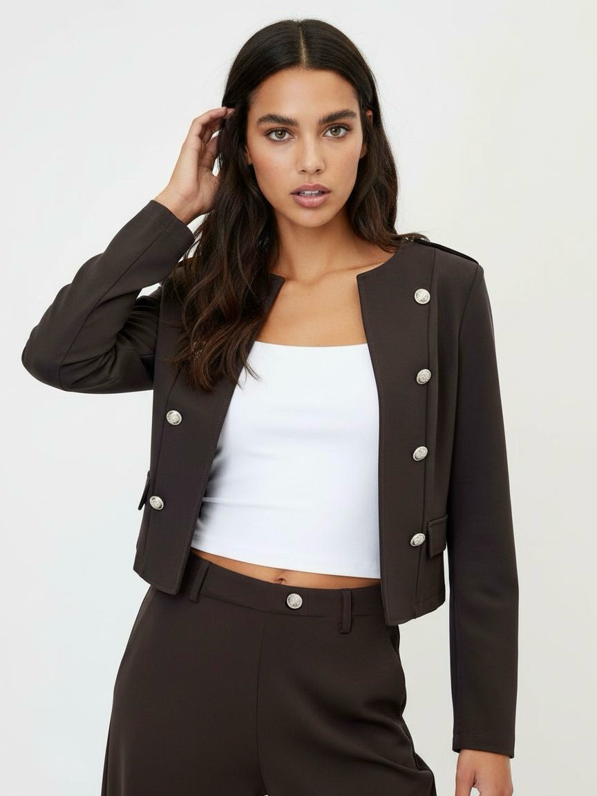 A woman modeling a dark brown cropped double-breasted blazer with silver buttons, paired with matching dark brown trousers and a white crop top, against a plain light gray background.