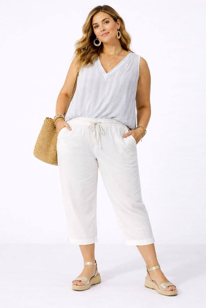 Woman wearing a white sleeveless top and white pants, holding a straw bag on a white background