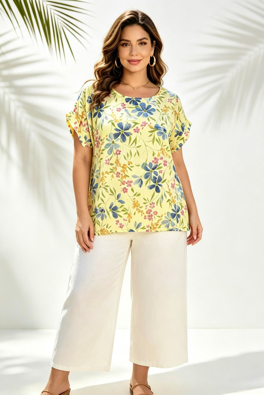 A woman modeling a short-sleeve yellow top with a blue, green, and pink floral print, paired with white cropped pants, set against a white background with palm leaf shadows.