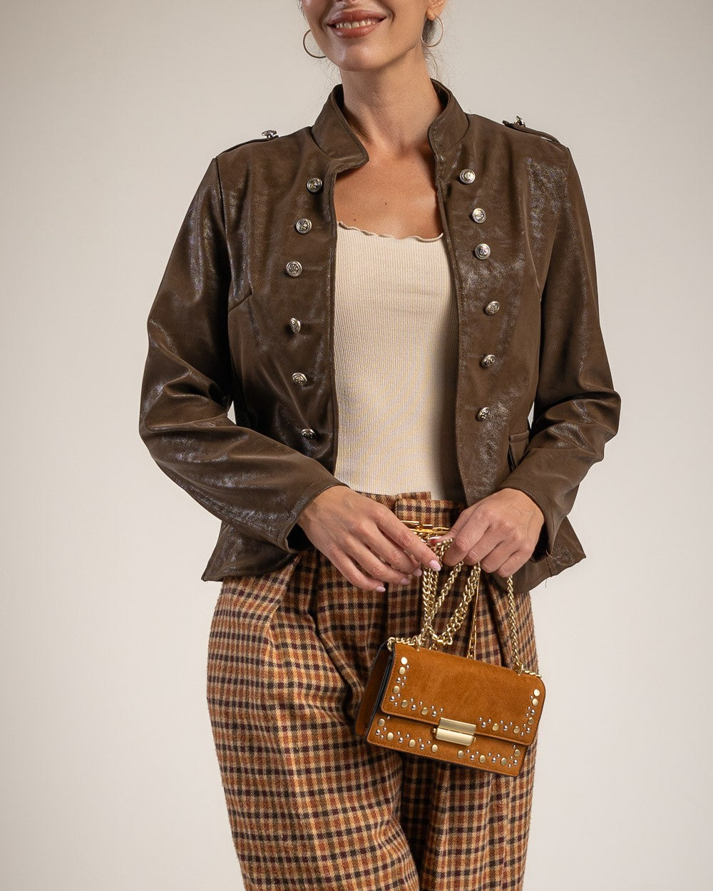 A woman wearing a brown faux-leather military-style jacket with silver buttons, paired with brown and beige plaid trousers and holding a small brown studded handbag, against a light beige studio background.