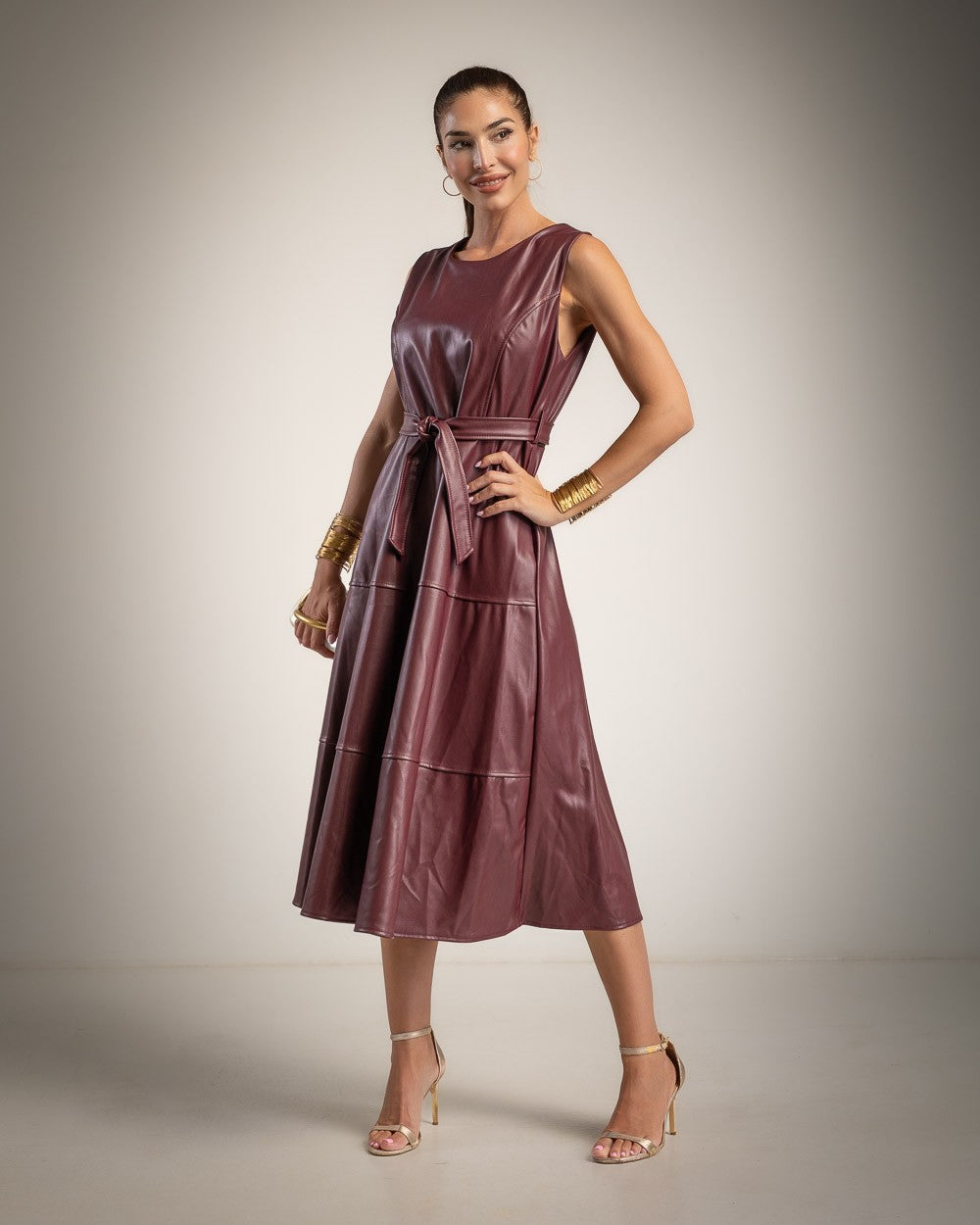 A woman models a sleeveless, tiered midi dress in a deep burgundy faux leather material, styled with gold bracelets, earrings, and nude high-heeled sandals against a neutral studio backdrop.